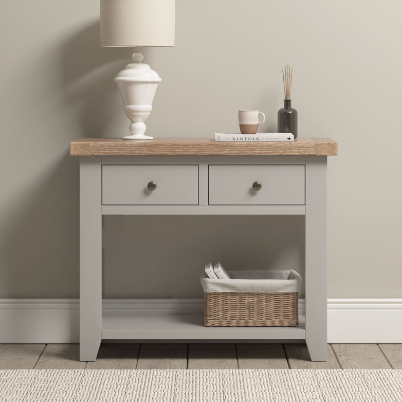 A Burford 2 Drawers Console Table in Pebble Grey with a lamp, cup, book, diffuser on top and a basket on the lower shelf.
