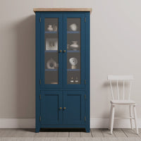 The Burford Display Cabinet in Navy Blue with glass doors displays decor, standing next to a white wooden chair against a light wall.