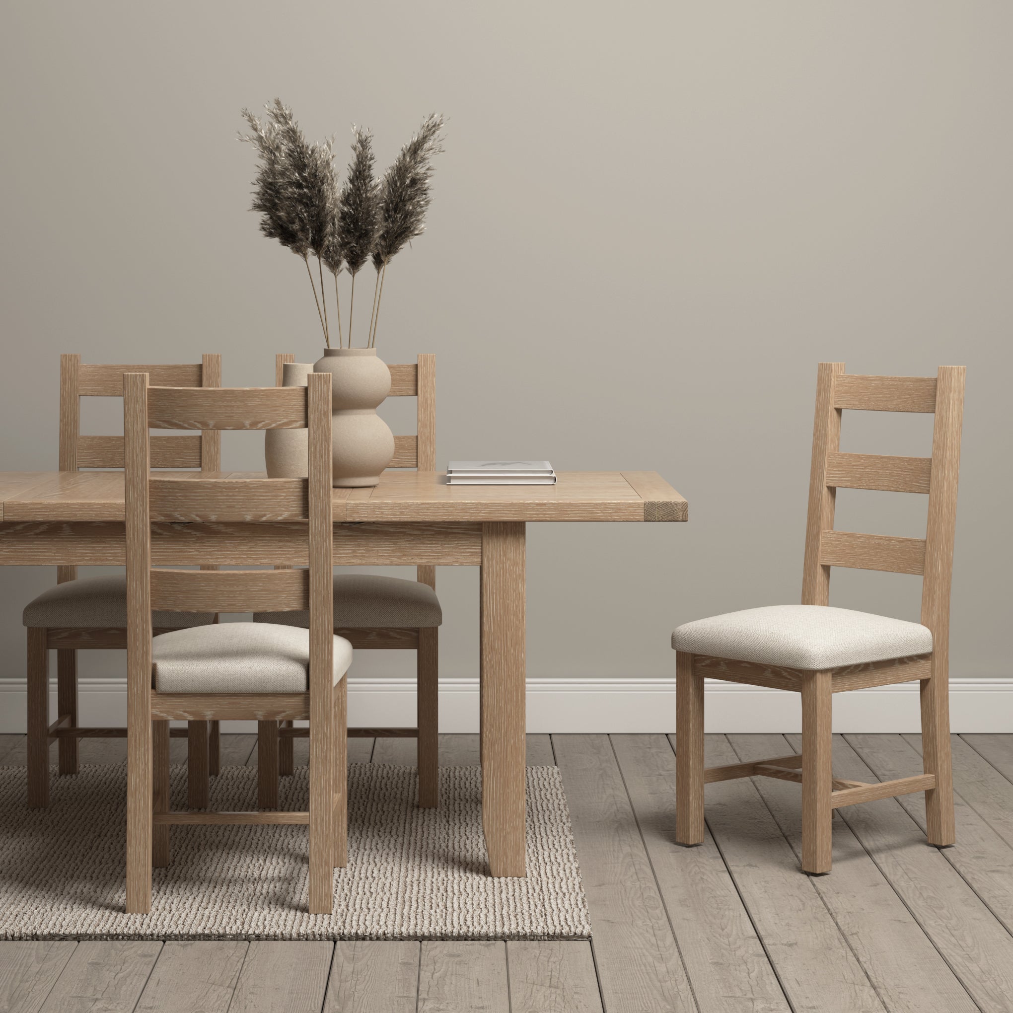 The minimalist dining room includes a Burford 6 Seat Extending Dining Set in Natural Oak and four chairs, with pampas grass in vases and books on a woven rug. Neutral tones blend seamlessly, creating an inviting ambiance.