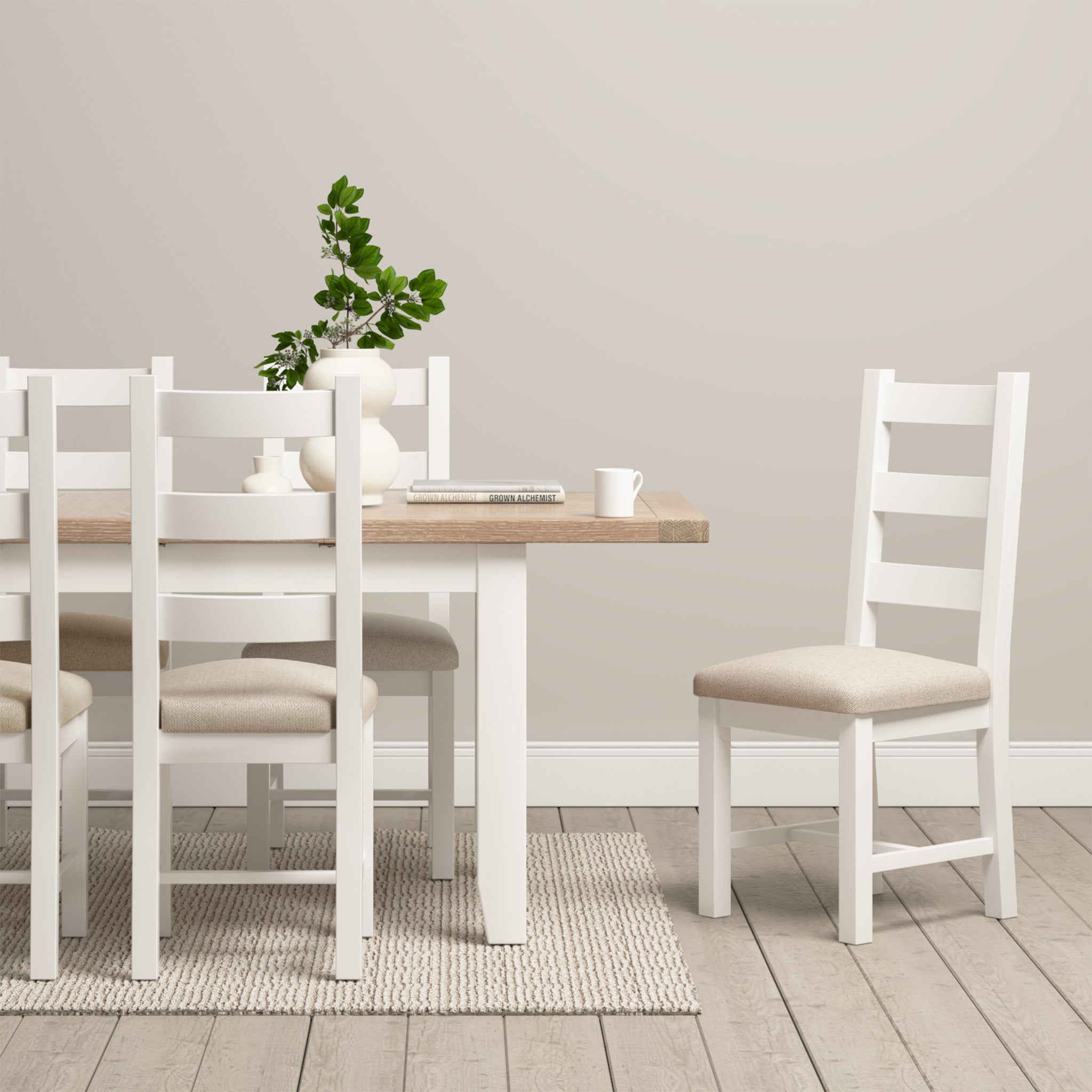 The minimalist dining room showcases the Burford 6 Seat Extending Dining Set in Warm White, complemented by surrounding white chairs. At its center is a plant centerpiece placed on a beige rug.