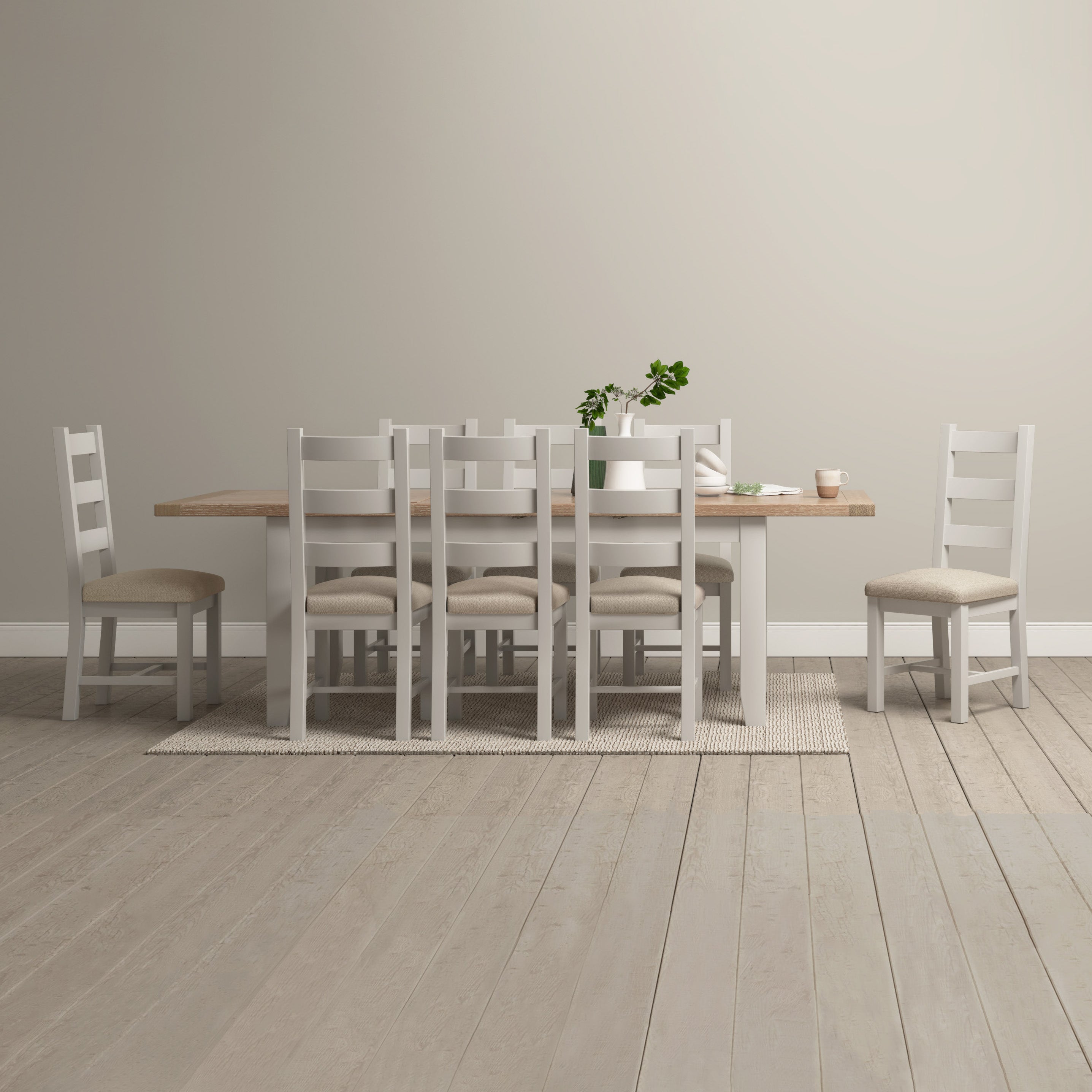 A Burford 8 Seat Extending Dining Set in Pebble Grey, featuring a wooden table and eight white chairs, is arranged with a small plant and dishes on a beige rug in a minimalist room.