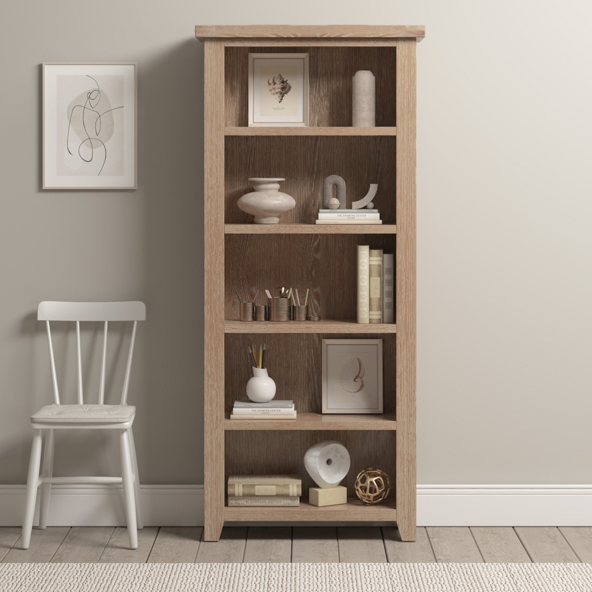 The Burford Large Bookcase in Natural Oak is styled with decor and books, set next to a white chair and framed wall art in a neutral-toned room.