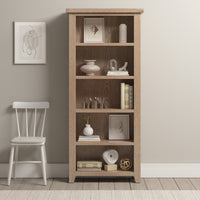 The Burford Large Bookcase in Natural Oak is styled with decor and books, set next to a white chair and framed wall art in a neutral-toned room.