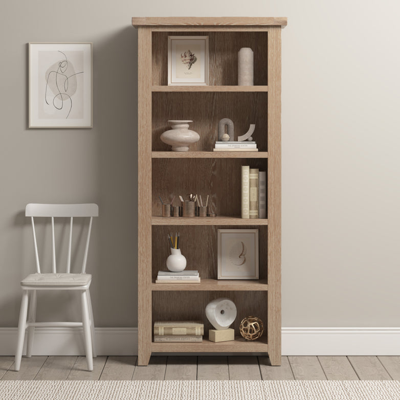 The Burford Large Bookcase in Natural Oak is styled with decor and books, set next to a white chair and framed wall art in a neutral-toned room.