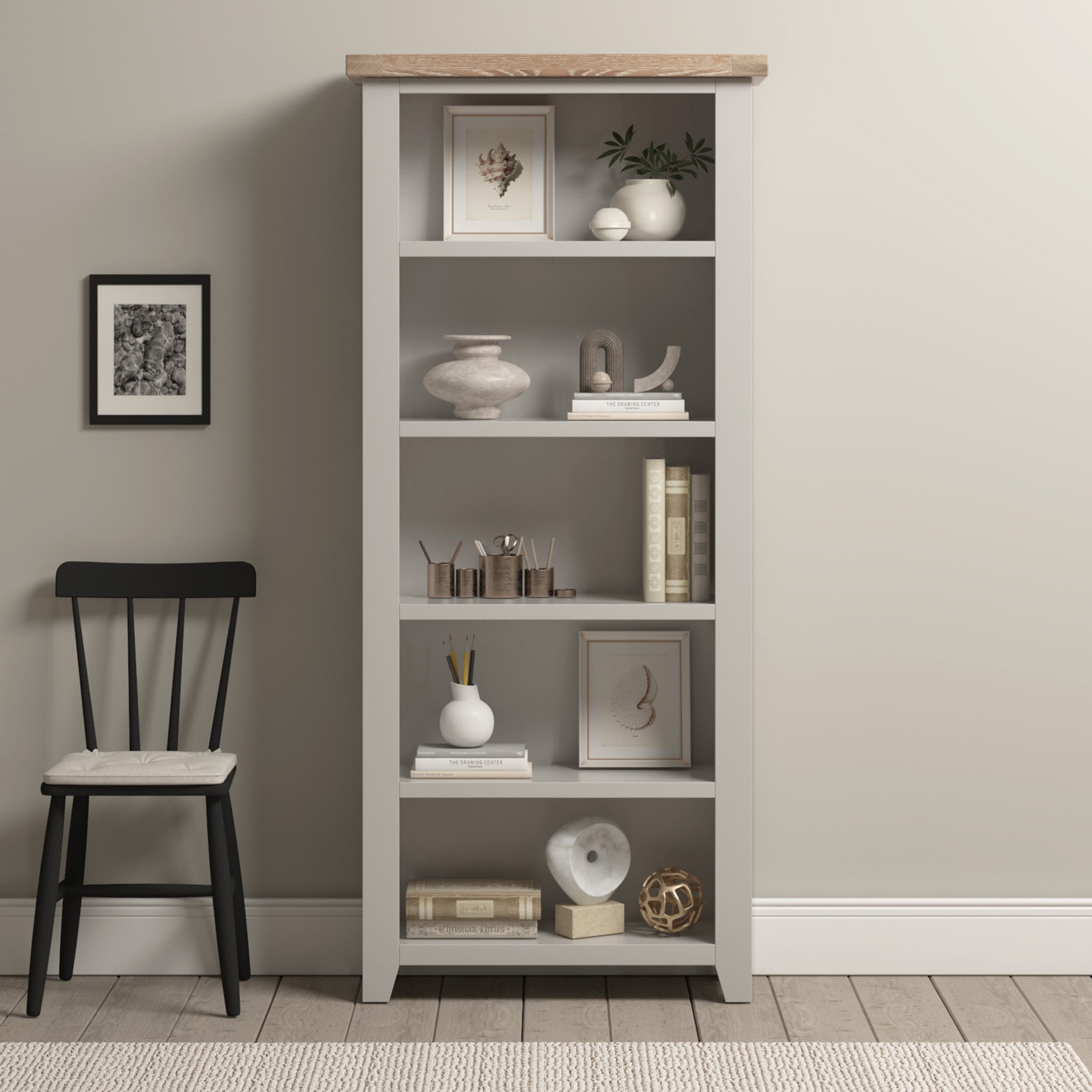 Burford Large Bookcase in Pebble Grey styled with decor, books, and art stands beside a black chair with a cushion against a beige wall.