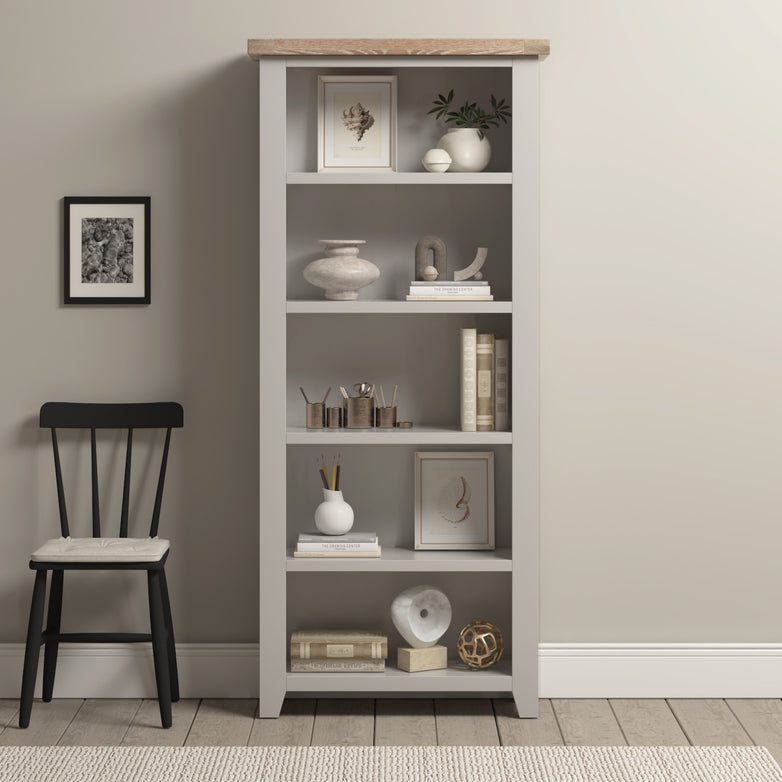 Burford Large Bookcase in Pebble Grey styled with decor, books, and art stands beside a black chair with a cushion against a beige wall.
