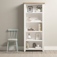 The Burford Large Bookcase in Warm White elegantly displays decor and books beside a light blue chair, harmonizing with the room’s beige walls and carpet, while its natural oak accents add sophistication.
