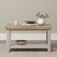 A Burford Large Coffee Table with Shelf in Pebble Grey, topped with books, a mug, a bowl, and a vase holding green branches.