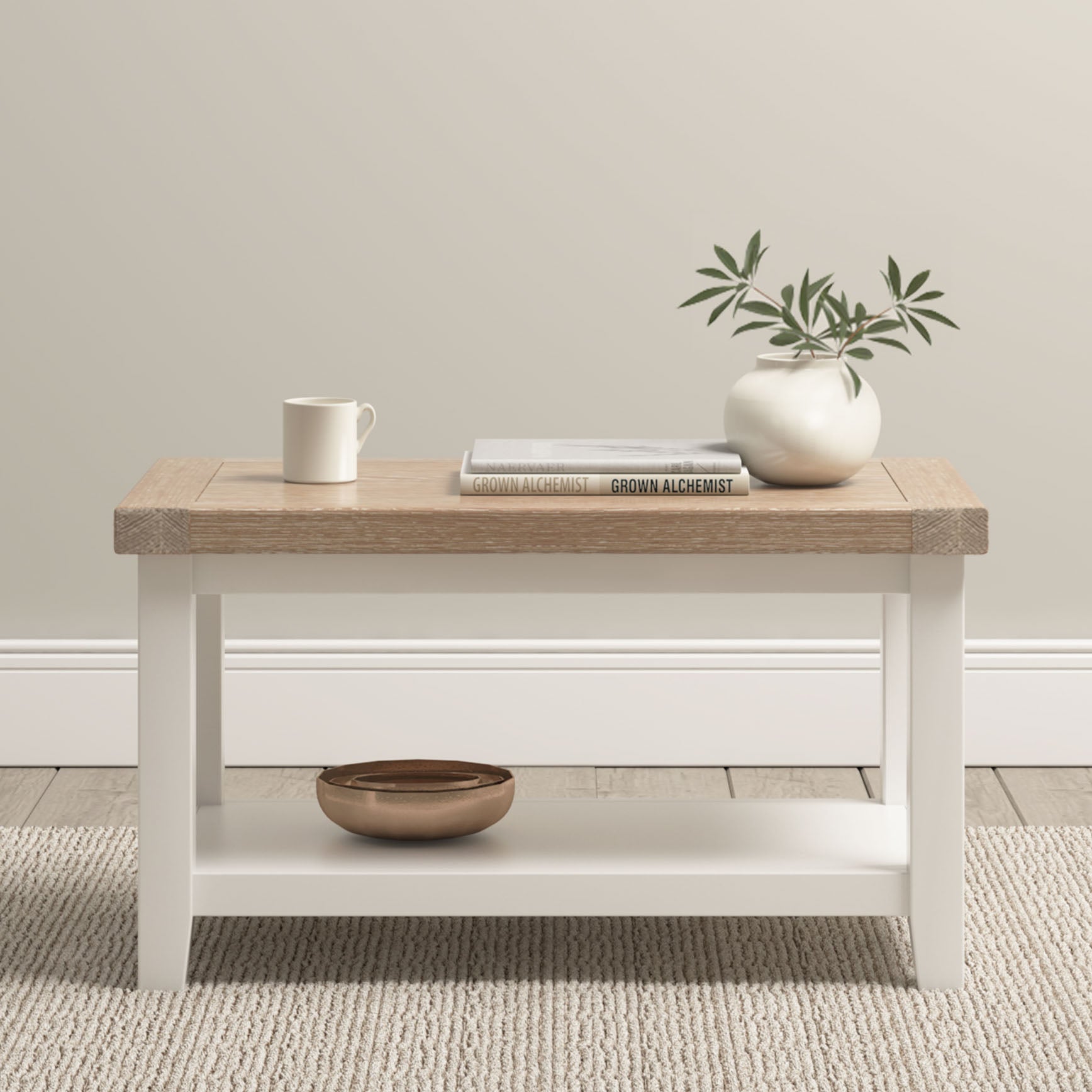 The Burford Large Coffee Table with Shelf in Warm White is styled with a mug, book, vase of greenery, and bowl, and sits on a light rug.