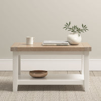 The Burford Large Coffee Table with Shelf in Warm White is styled with a mug, book, vase of greenery, and bowl, and sits on a light rug.