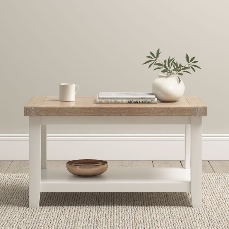 The Burford Large Coffee Table with Shelf in Warm White is styled with a mug, book, vase of greenery, and bowl, and sits on a light rug.