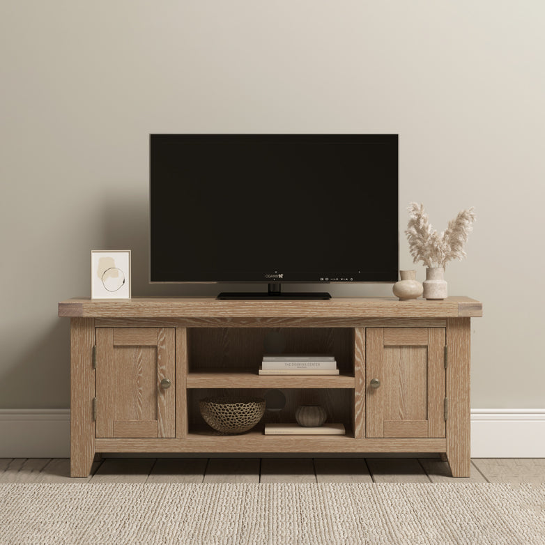 A flat-screen TV sits on the Burford 120cm Media Unit in Natural Oak with decor items, set against a beige wall atop a textured rug.