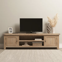 The Burford Large 180cm Media Unit in Natural Oak is a stylish wooden TV stand, ideal for living room storage and display of a flat-screen TV, vase with pampas grass, photo frame, and decorative accessories.