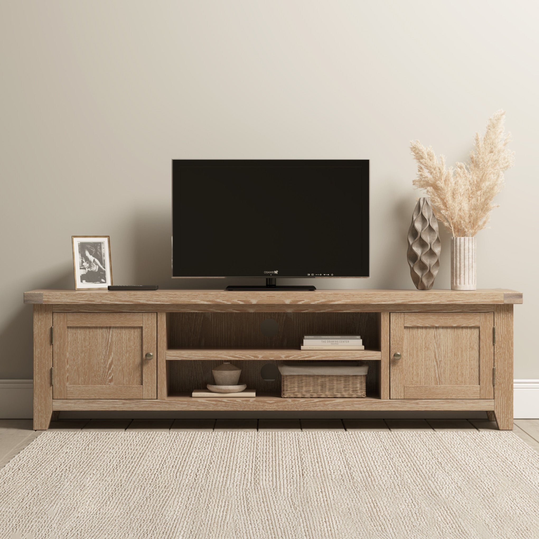 The Burford Large 180cm Media Unit in Natural Oak is a stylish wooden TV stand, ideal for living room storage and display of a flat-screen TV, vase with pampas grass, photo frame, and decorative accessories.