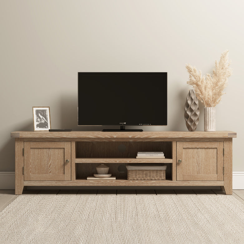 The Burford Large 180cm Media Unit in Natural Oak is a stylish wooden TV stand, ideal for living room storage and display of a flat-screen TV, vase with pampas grass, photo frame, and decorative accessories.