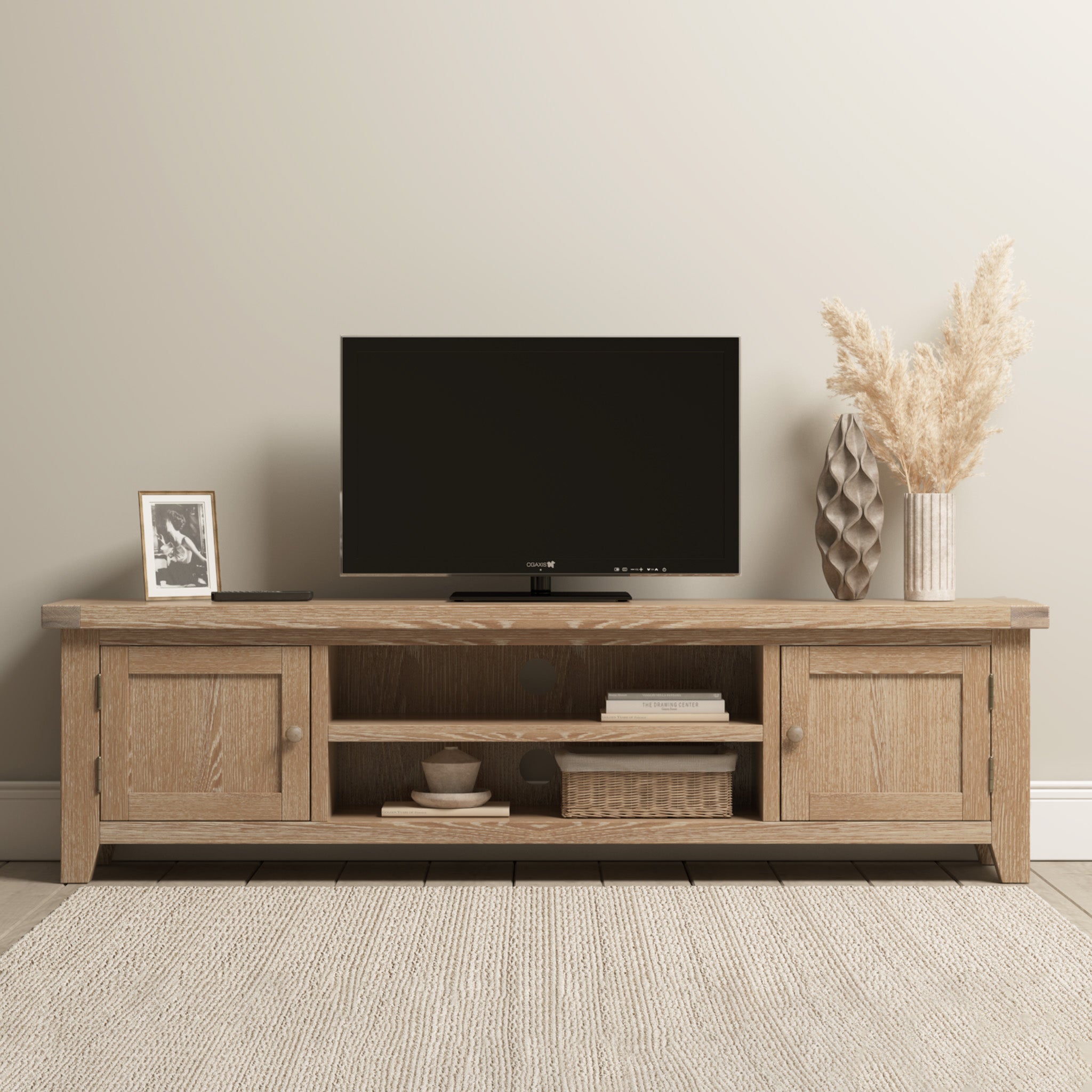 The Burford Large 180cm Media Unit in Natural Oak is styled with a TV, photo frame, vase of dried grasses, and woven basket on a light rug.