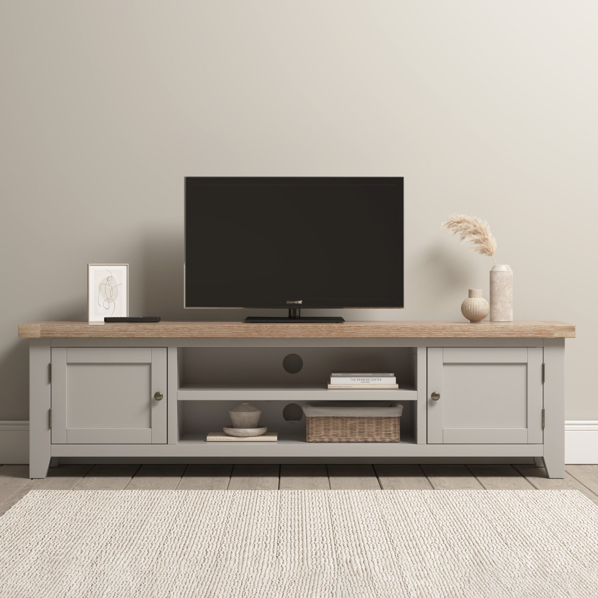 The Burford Large 180cm Media Unit in Pebble Grey showcases a flat-screen TV, decor, and baskets on a light rug by a beige wall. Nearby, a Burford Ladderback Dining Chair with a fabric seat adds comfort and style.