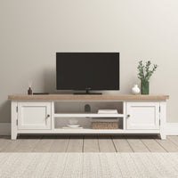 A TV sits on the Burford Large 180cm Media Unit in Warm White, adorned with decor items like a vase and basket in a bright, minimalist room. Nearby, the Burford Ladderback Dining Chair with a fabric seat and natural oak finish enhances the cozy yet stylish ambiance.