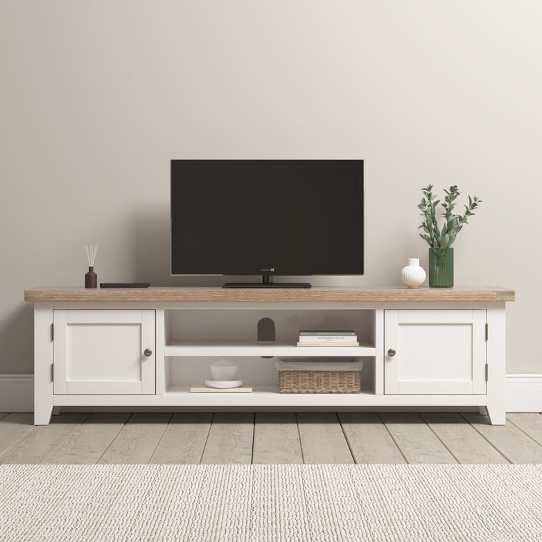 A TV sits on the Burford Large 180cm Media Unit in Warm White, adorned with decor items like a vase and basket in a bright, minimalist room. Nearby, the Burford Ladderback Dining Chair with a fabric seat and natural oak finish enhances the cozy yet stylish ambiance.