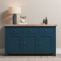 The Burford Large 3 Door 3 Drawers Sideboard in Navy Blue features a wooden top with a lamp, small vase with leaves, and two black bottles, offering ample storage and timeless style.