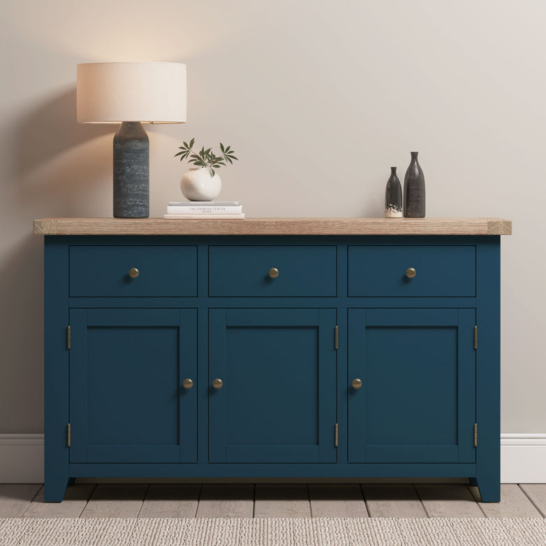 The Burford Large 3 Door 3 Drawers Sideboard in Navy Blue features a wooden top with a lamp, small vase with leaves, and two black bottles, offering ample storage and timeless style.