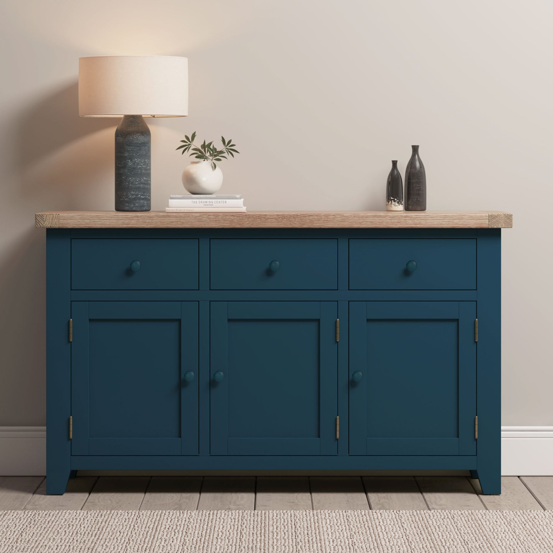 The Burford Large 3 Door 3 Drawers Sideboard in Navy Blue with a wooden top is styled with a lamp, books, small plant, and two vases against a beige wall.