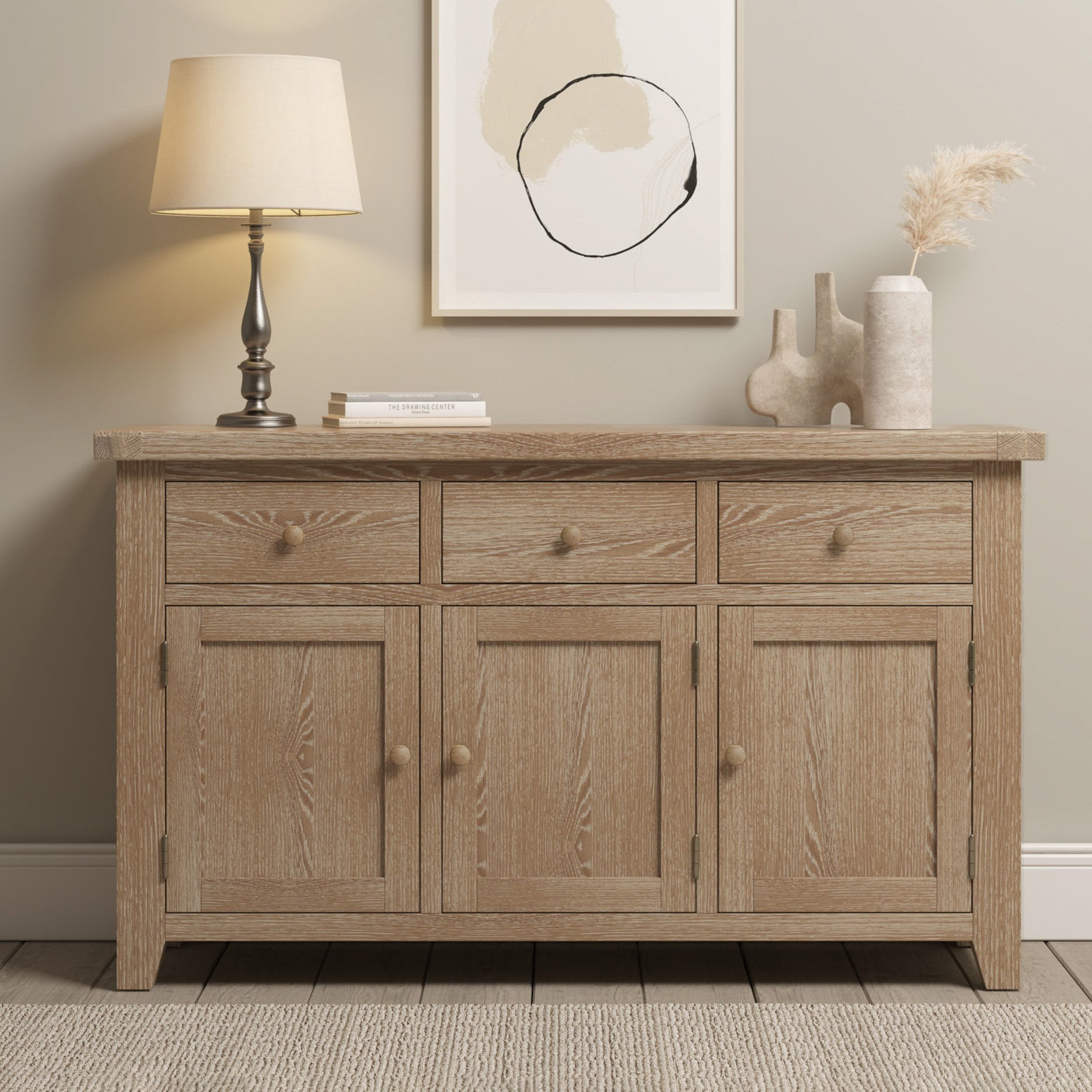 The Burford Large 3 Door 3 Drawers Sideboard in Natural Oak, topped with a lamp, books, and vases, sits beneath abstract wall art in a cozy room.