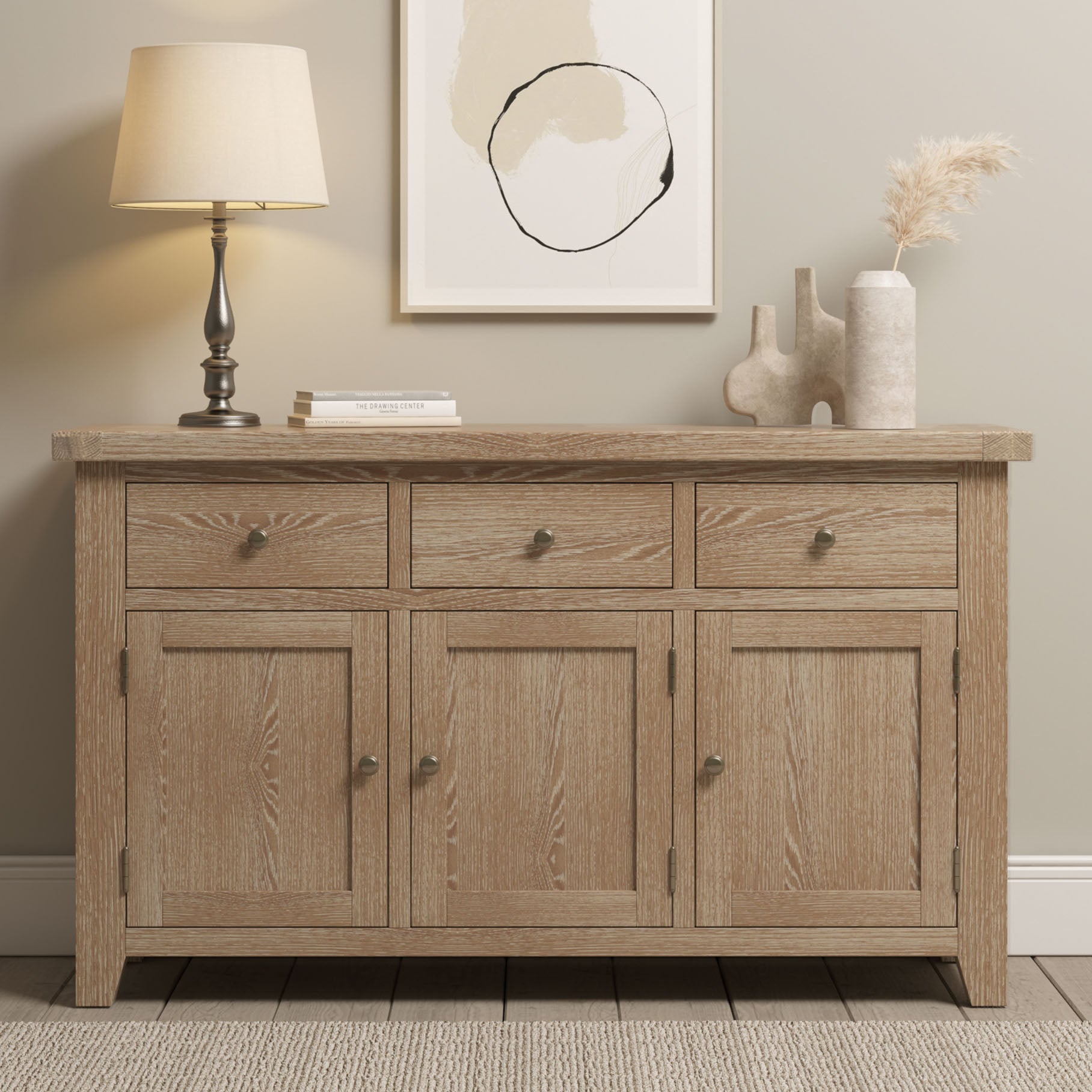 Natural oak Burford Large Sideboard with three drawers and three doors, styled with a lamp, vases, and abstract wall art above.