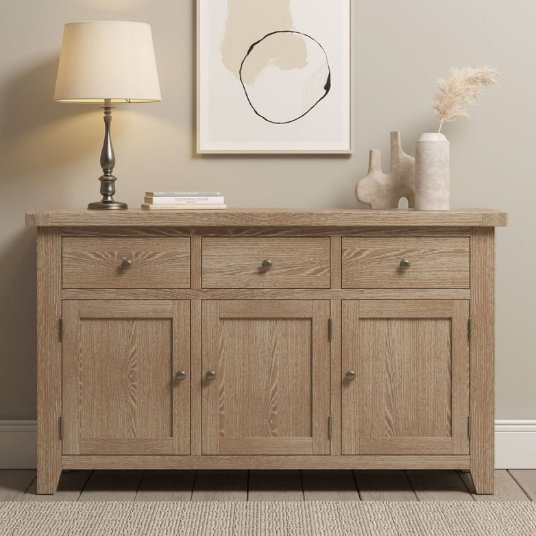 Natural oak Burford Large Sideboard with three drawers and three doors, styled with a lamp, vases, and abstract wall art above.