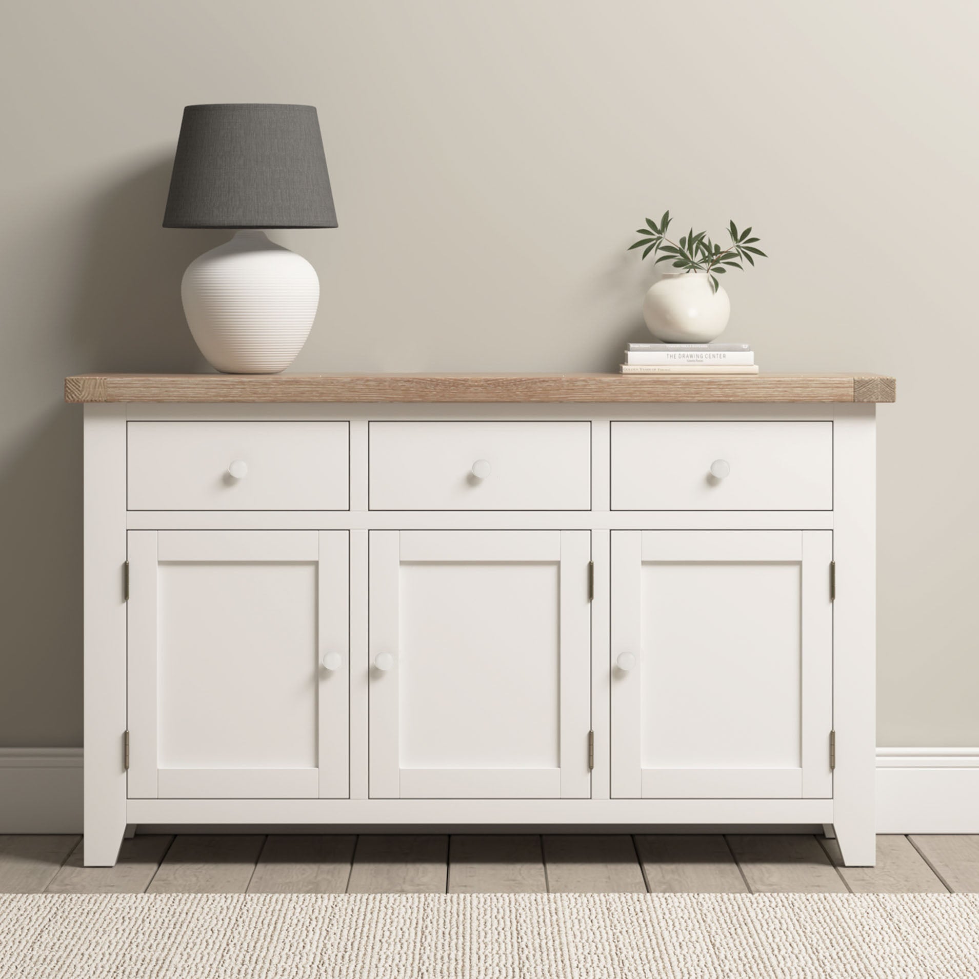 Burford Large 3 Door 3 Drawers Sideboard in Warm White with an oak top, styled with a lamp, small plant, and books against a beige wall.