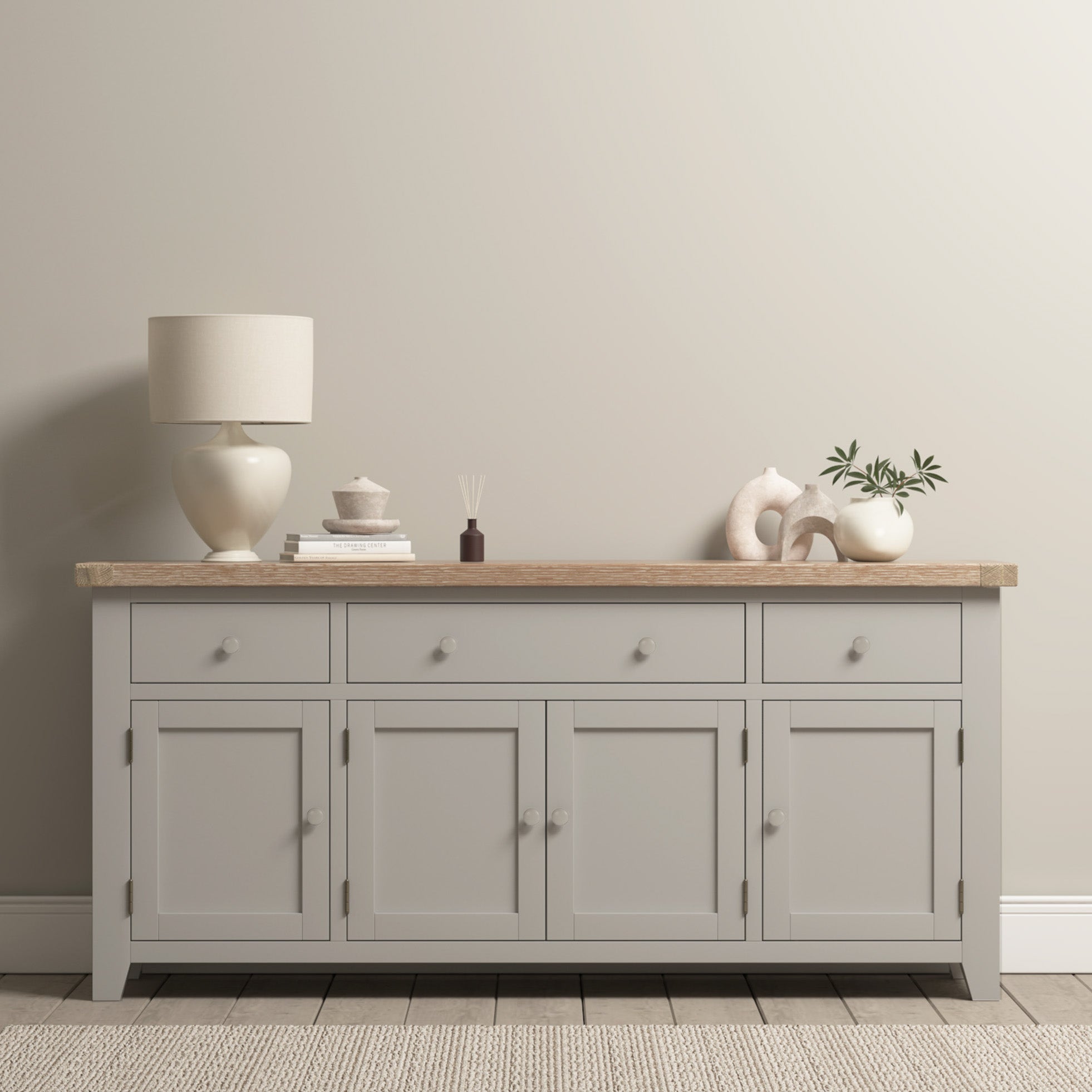 The Burford Large 4 Door 3 Drawers Sideboard in Pebble Grey is shown against a beige wall, topped with decor items, a lamp, books, and a plant.