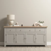 The Burford Large 4 Door 3 Drawers Sideboard in Pebble Grey is shown against a beige wall, topped with decor items, a lamp, books, and a plant.