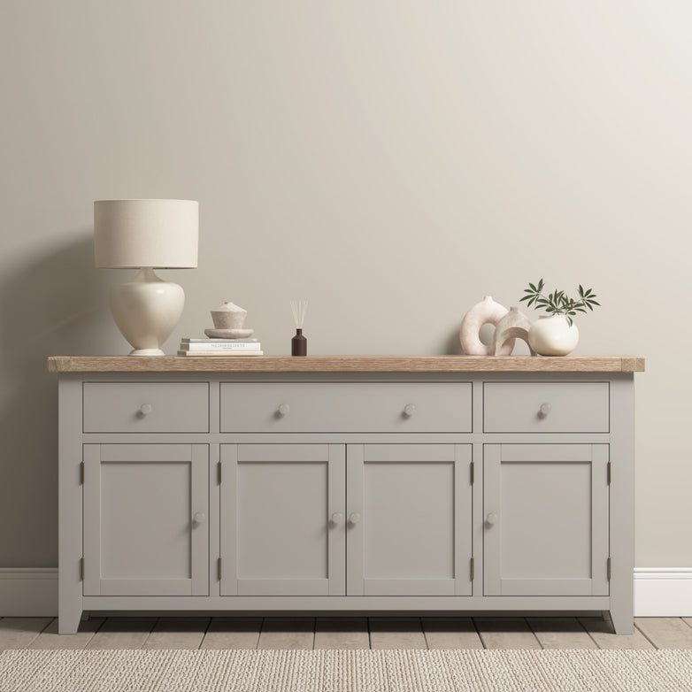 The Burford Large 4 Door 3 Drawers Sideboard in Pebble Grey is shown against a beige wall, topped with decor items, a lamp, books, and a plant.