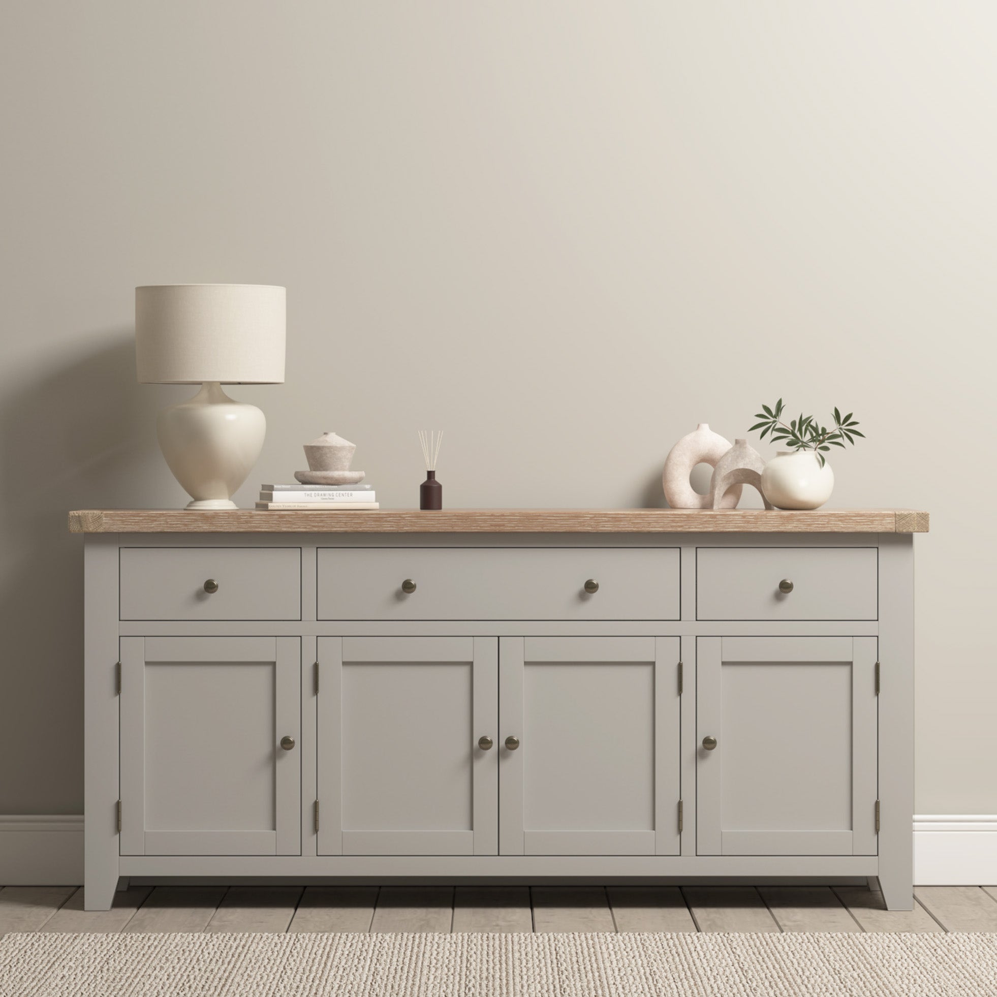 Burford Large 4 Door 3 Drawers Sideboard in Pebble Grey with a lamp, decor, and plants on top stands against a beige wall on a woven rug.
