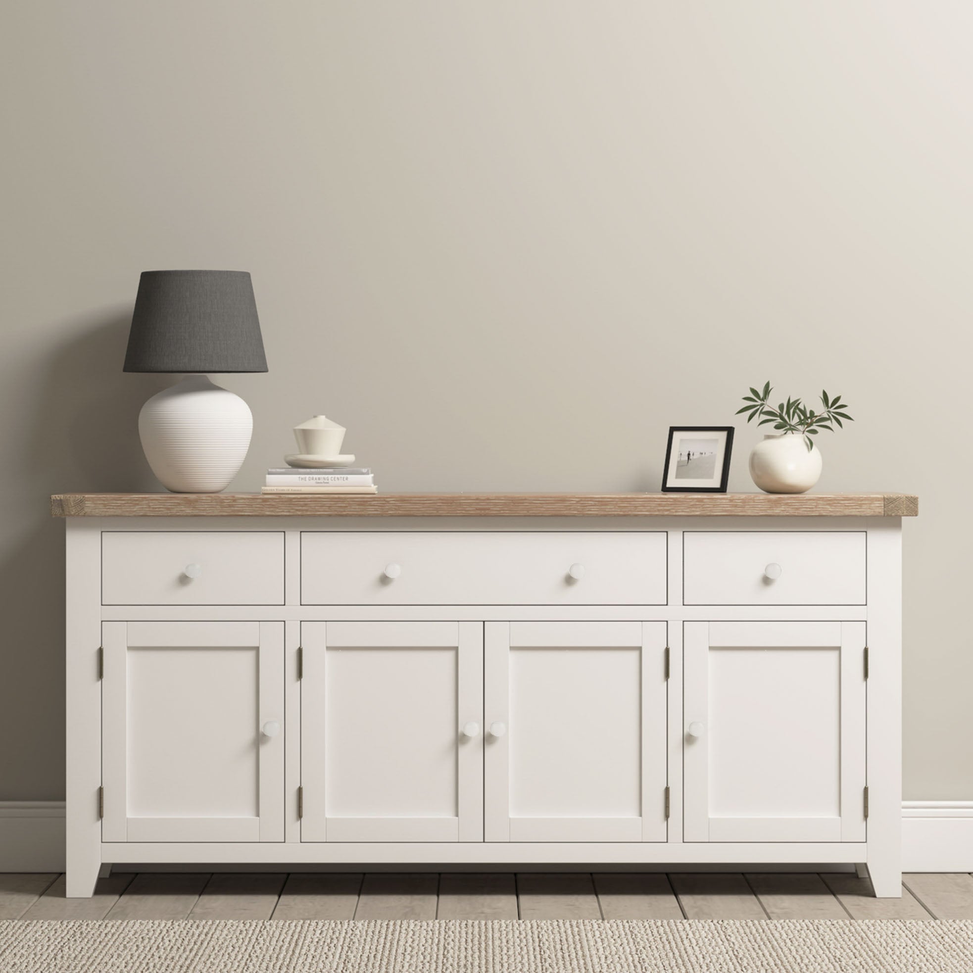 The Burford Large 4 Door 3 Drawers Sideboard in Warm White with a natural oak top is styled with a lamp, teacup, framed photo, and plant against a beige wall.