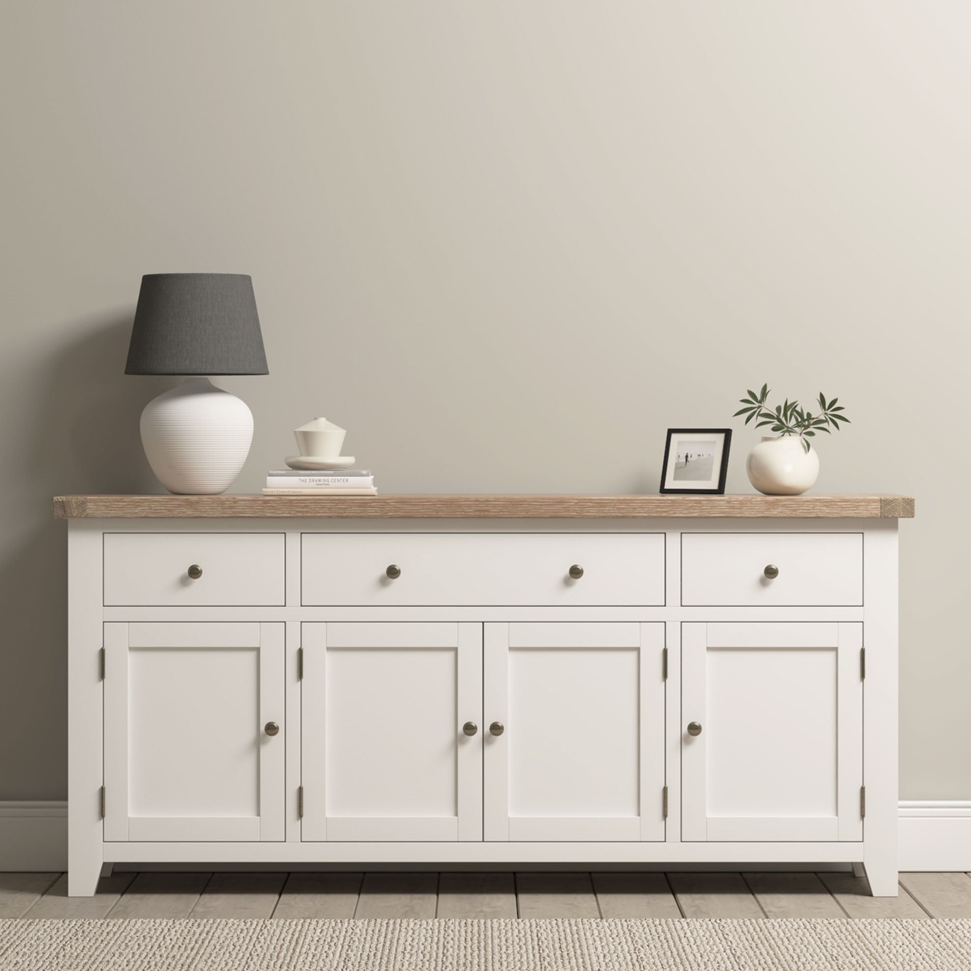 The Burford Large 4 Door 3 Drawers Sideboard in Warm White is topped with a lamp, framed photo, plant, and teacup, set against a neutral wall.