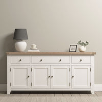 The Burford Large 4 Door 3 Drawers Sideboard in Warm White is topped with a lamp, framed photo, plant, and teacup, set against a neutral wall.
