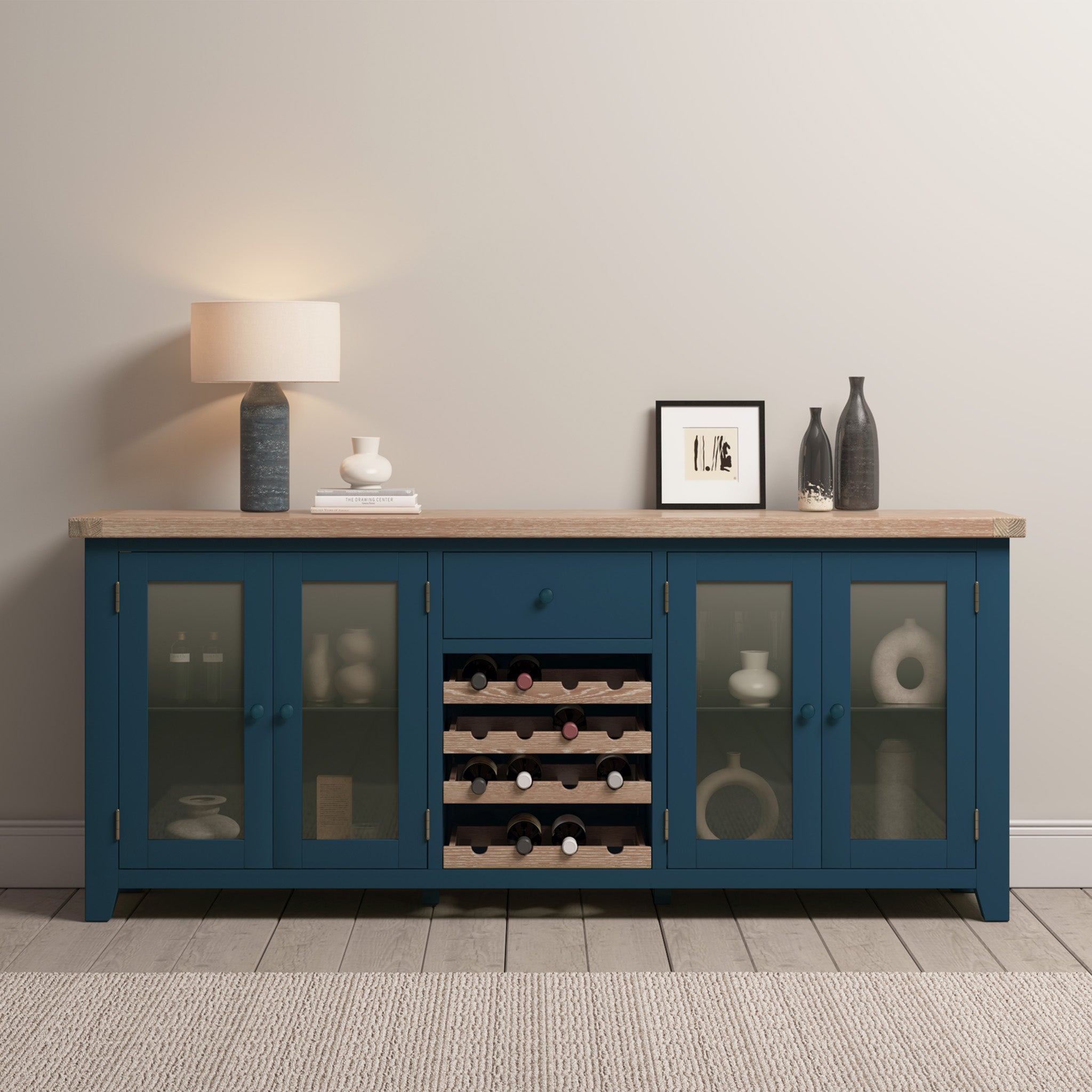 A Burford Grand Sideboard with Wine Rack in Navy Blue sits against a light wall on a wood floor, styled with glass doors, a lamp, and decor.