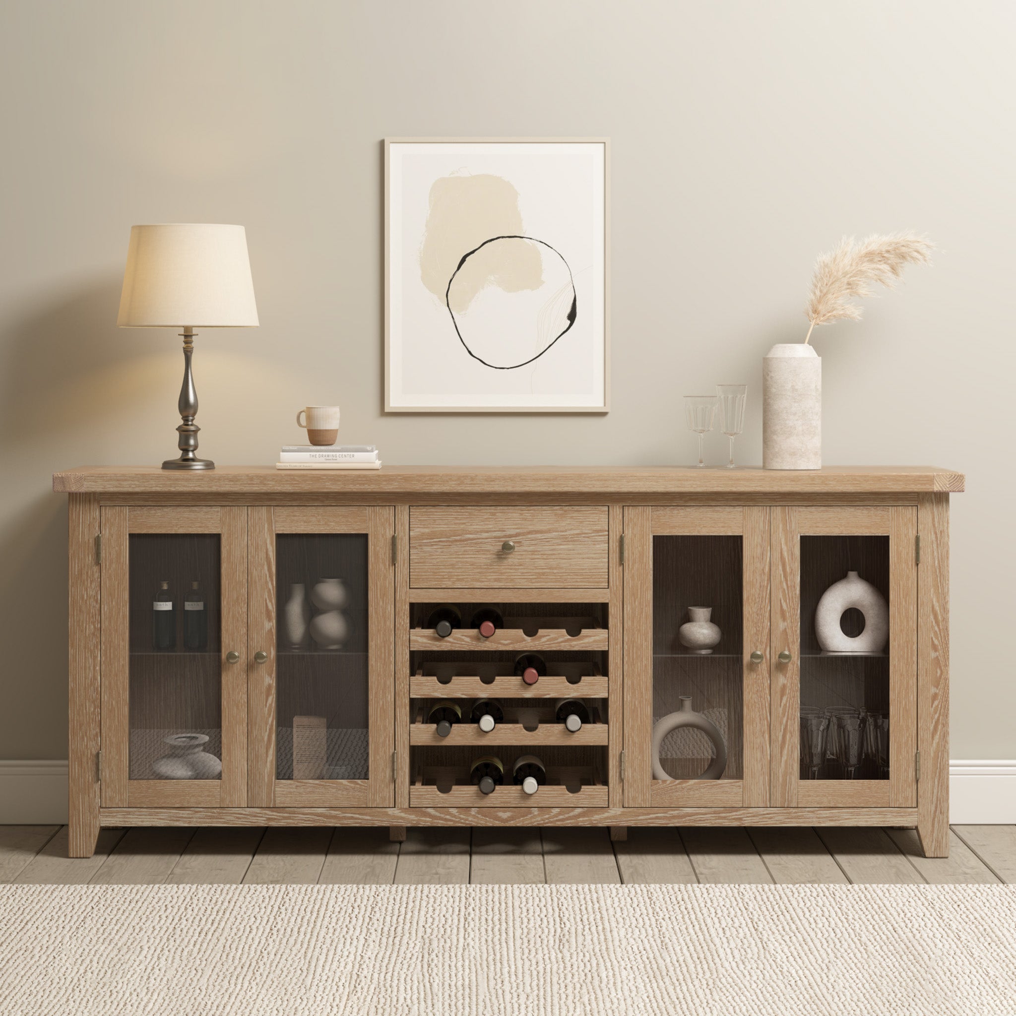 The Burford Grand Sideboard with Wine Rack in Natural Oak, paired with glass doors, a stylish lamp, and abstract wall art, brings modern flair to a neutral room.