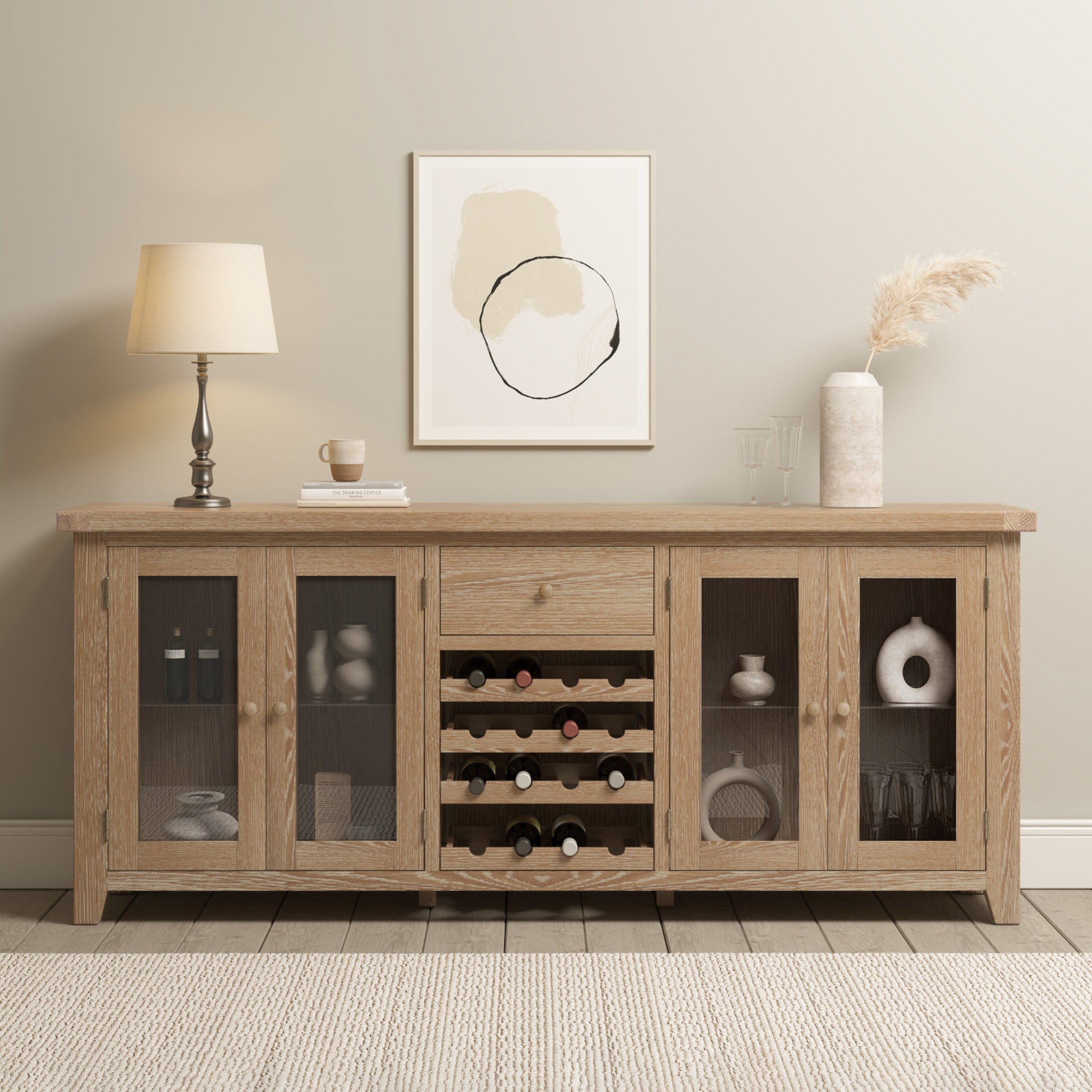 The Burford Grand Sideboard with Wine Rack in Natural Oak features glass cabinets and is styled with décor, a lamp, and abstract art, set against a beige wall.
