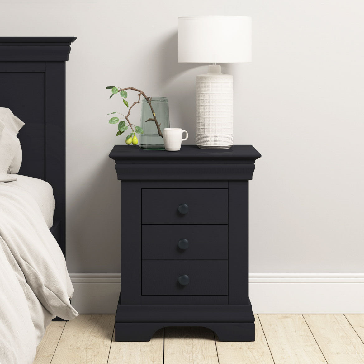 The Chalbury 3 Drawer Bedside Table in Dusky Black provides ample bedroom storage with three drawers and sits neatly beside the bed, topped with a white lamp, glass jug, cup, and a small plant.