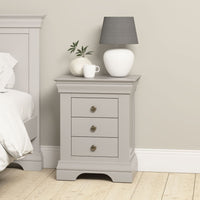 The Chalbury 3 Drawer Bedside Table in Pebble Grey stands beside a bed, topped with a white lamp, a white mug, and a small plant.