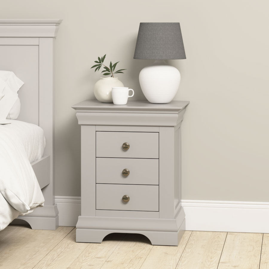 The Chalbury 3 Drawer Bedside Table in Pebble Grey stands beside a bed, topped with a white lamp, a white mug, and a small plant.