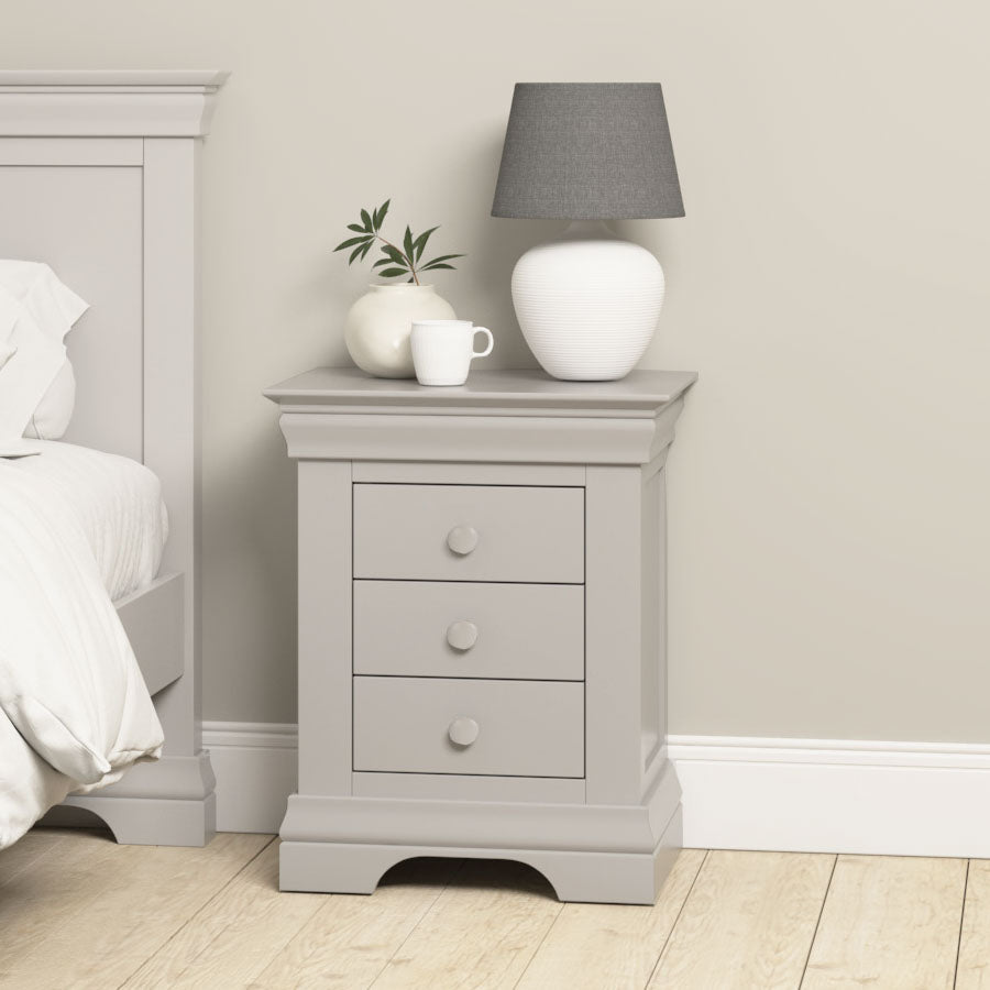 The Chalbury 3 Drawer Bedside Table in Pebble Grey is shown beside a bed on a light wood floor, paired with a white lamp, plant, and mug.