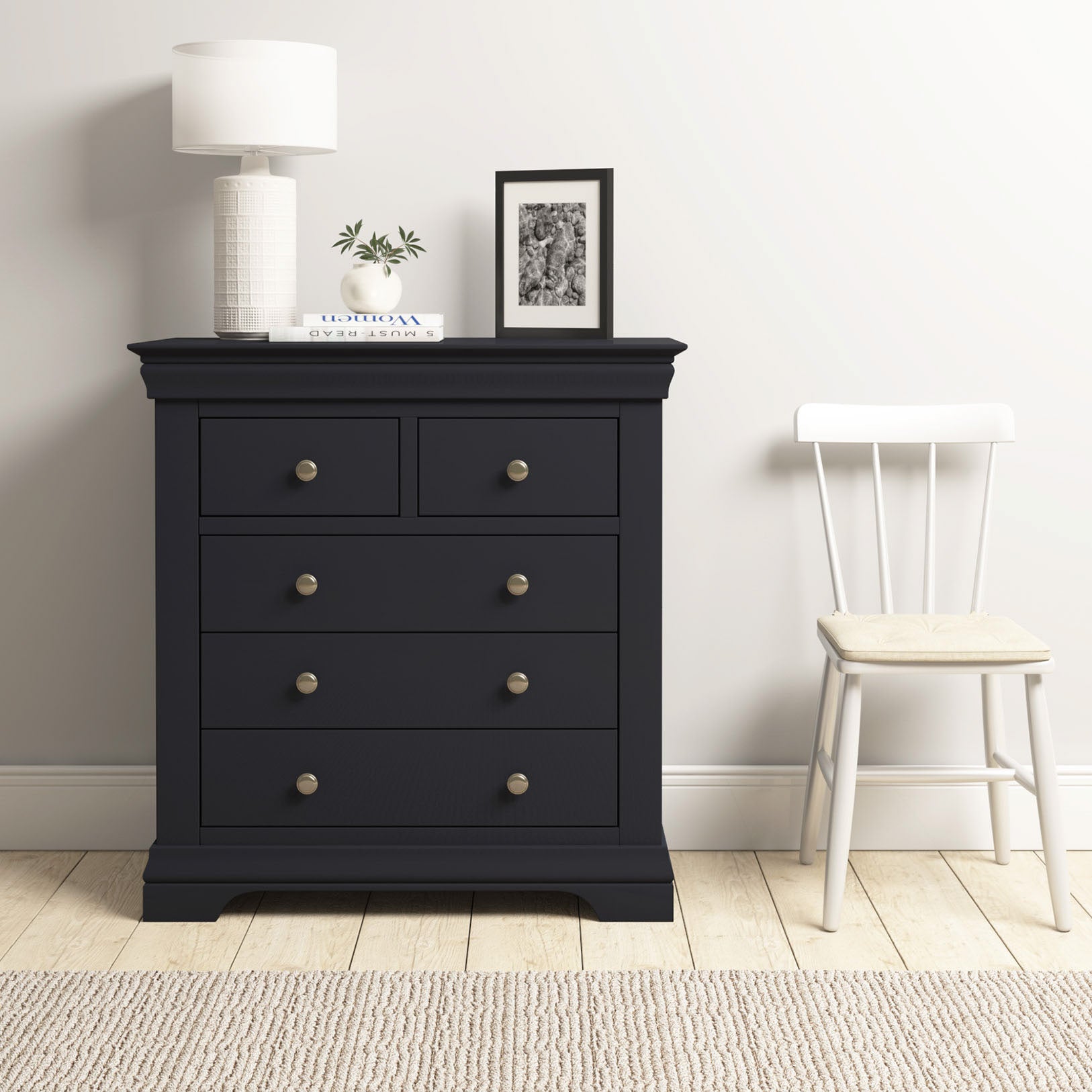 The Chalbury 2 Over 3 Chest of Drawers in Dusky Black, topped with a lamp, plant, and photo, stands beside a white chair in a minimalist room.