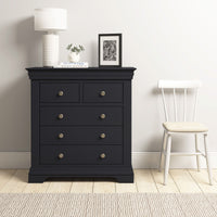 The Chalbury 2 Over 3 Chest of Drawers in Dusky Black, topped with a lamp, plant, and photo, stands beside a white chair in a minimalist room.