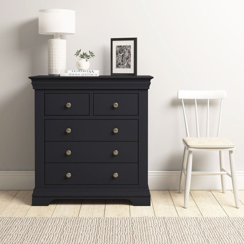 The Chalbury 2 Over 3 Chest of Drawers in Dusky Black, topped with a lamp, plant, and photo, stands beside a white chair in a minimalist room.
