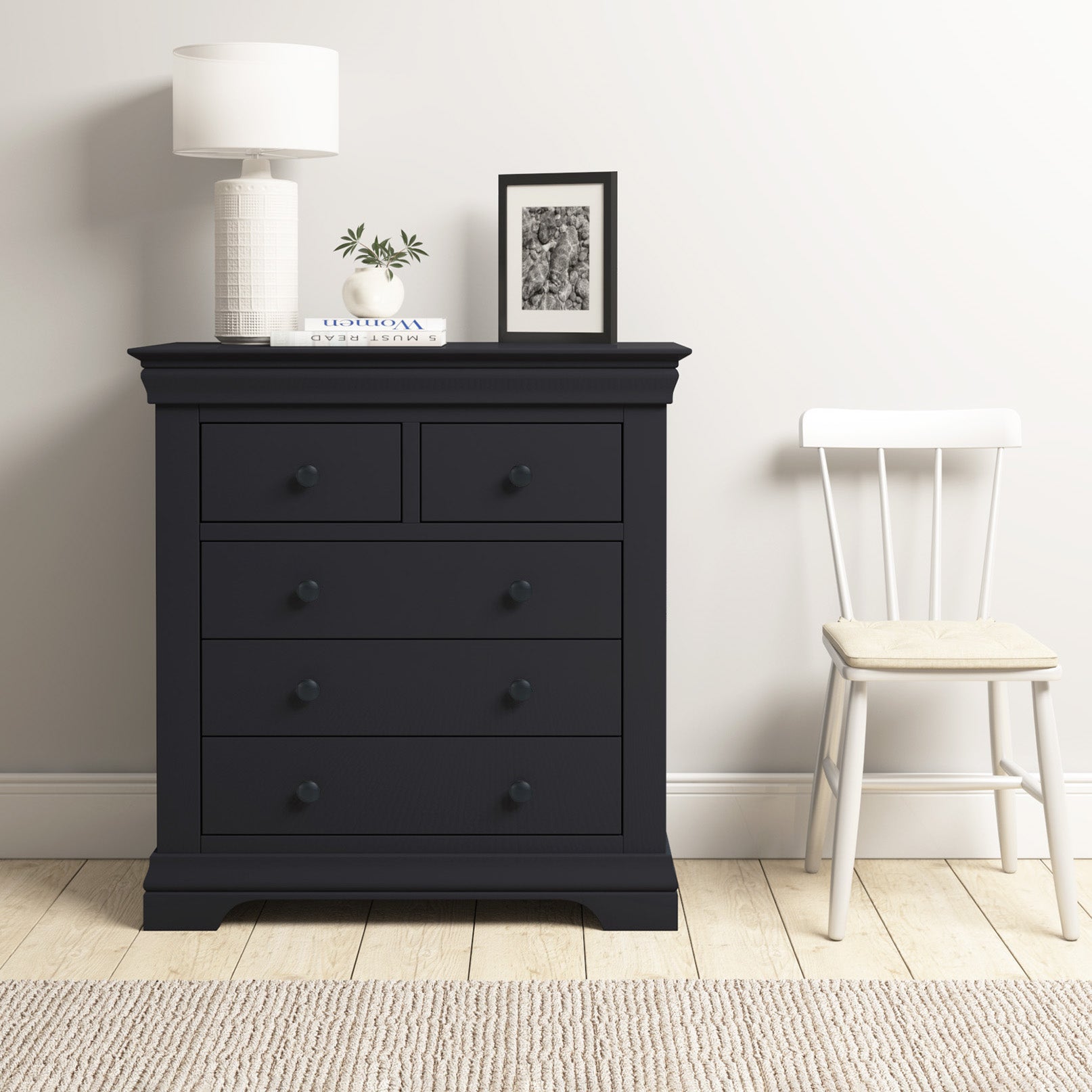 A Chalbury 2 Over 3 Chest of Drawers in Dusky Black holds a lamp, books, plant, and framed photo next to a white chair in this bright, modern room.