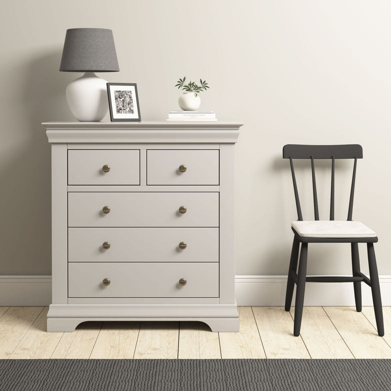 The Chalbury 2 Over 3 Chest of Drawers in Pebble Grey sits beside a black chair, styled with a lamp, photo, and plant for a modern touch.