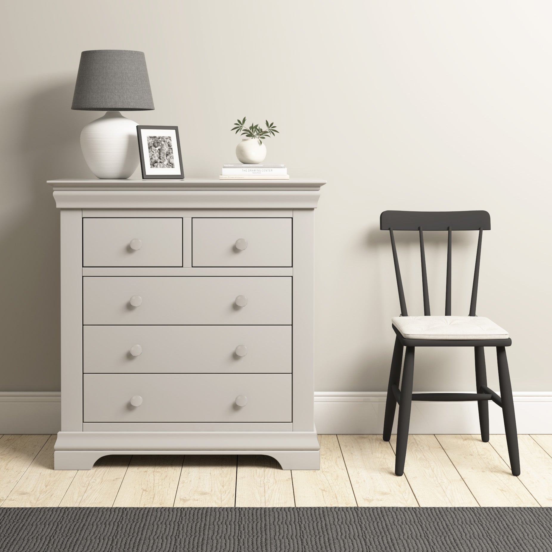 The Chalbury 2 Over 3 Chest of Drawers in Pebble Grey is styled with decor, placed next to a black chair on a wooden floor in a minimalist room.